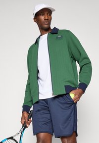 Casaco verde com fecho éclair, com colarinho e punhos em azul-marinho, usado sobre uma camisa branca. Calções azul-marinho, segurando uma bola de ténis e uma raquete.