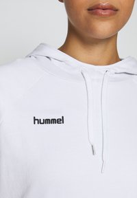 Gros plan d'une personne portant un sweat à capuche blanc avec des cordons blancs et un logo noir "hummel" sur la poitrine.