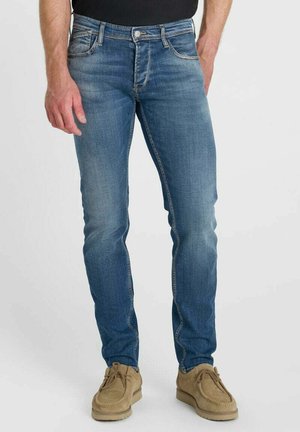 Man wearing slim-fit blue jeans and tan suede shoes, standing against a plain light background.