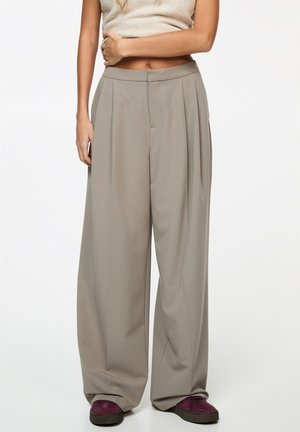 Vrouw die beige broek met wijde pijpen draagt, mouwloos beige topje en bordeauxrode schoenen, met één arm gekruist over haar buik.