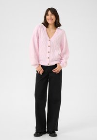 Cardigan en tricot rose clair avec un col en V, motif texturé et six boutons en bois. Associé à un pantalon large noir et des chaussures.