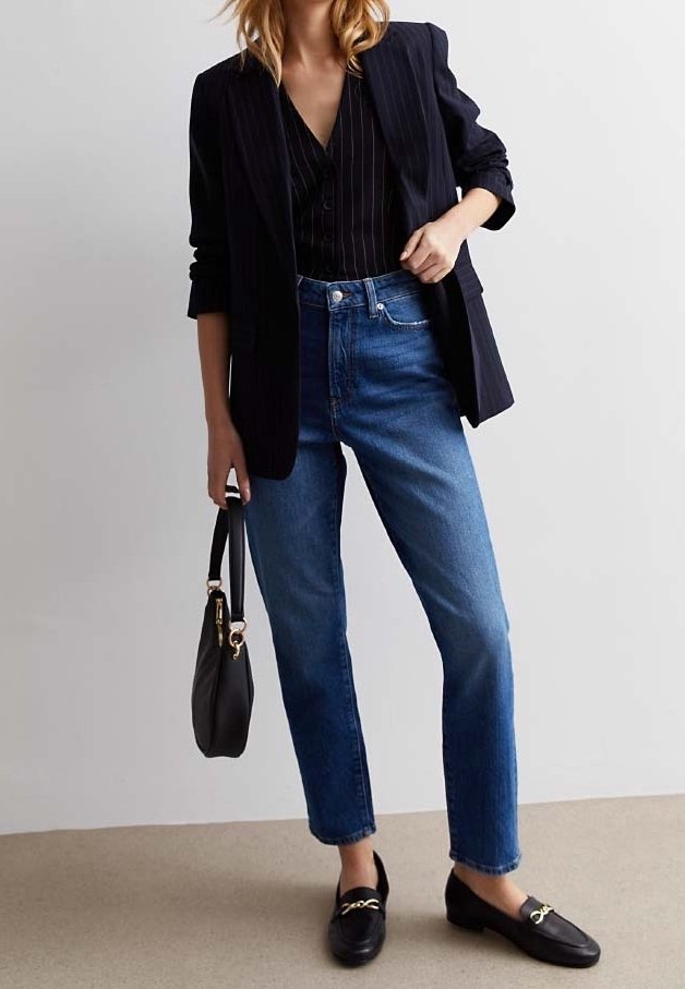 Femme vêtue d'un blazer à fines rayures sombres, blouse noire, jean taille haute bleu, mocassins noirs, tenant un sac à main noir à ses côtés.