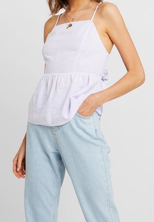 Woman wearing a white sleeveless peplum top with thin straps and light blue high-waisted jeans, hand in pocket, against a plain background.