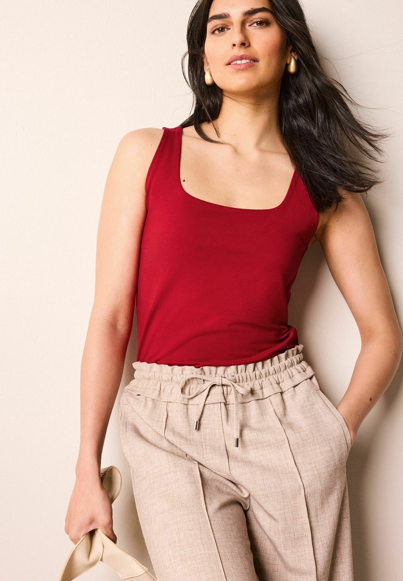 Top sin mangas de cuello redondo en rojo combinado con pantalones beige texturizados que tienen una cintura elástica y cordón. Modelo sosteniendo un bolso de color claro.