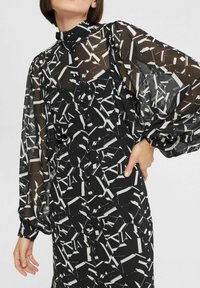 Schwarz-weiße gemusterte Bluse mit durchsichtigen langen Ärmeln, hohem Kragen und durchgehender Knopfleiste. Der Stoff hat ein leichtes, strukturiertes Aussehen.