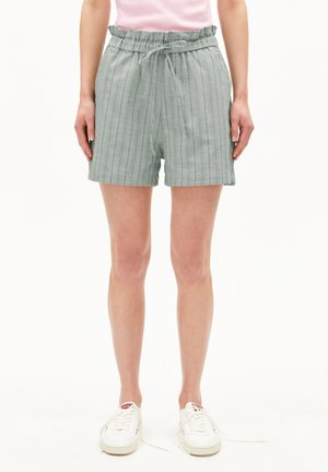 Personne portant un short à carreaux gris clair avec une taille à cordon, une chemise rose rentrée et des baskets blanches aux lacets défaits.