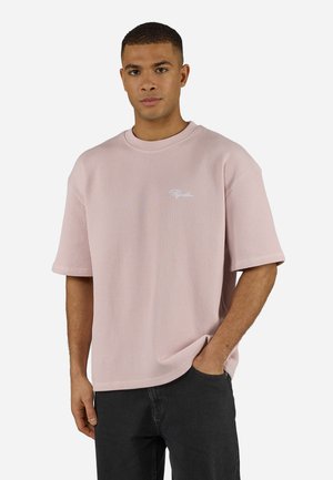 Mann iført lys rosa ribbestrikket kortermet skjorte med liten hvit logo på brystet, svarte bukser, stående med en hånd i lomma.