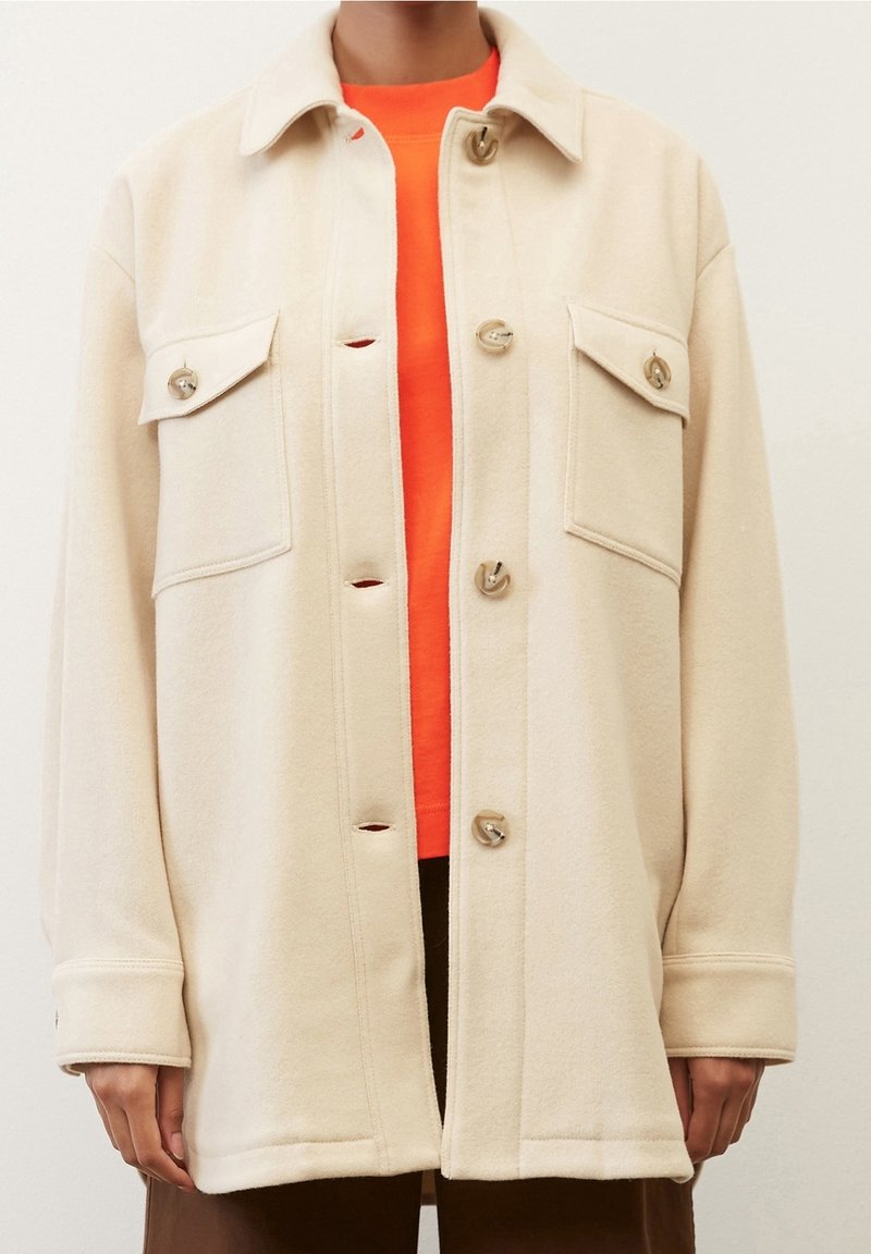 Veste beige surdimensionnée à boutons avec deux poches à rabat sur la poitrine, portée sur un haut orange vif et un pantalon marron.
