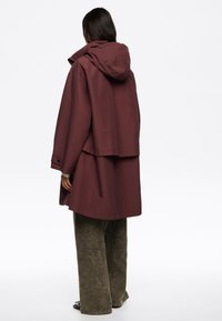 Bordo dežni jopič s kapuco, oversize oblikovanja in zategnjenimi rokavi. V kombinaciji z teksturiranimi širokimi olivno zelenimi hlačami, ki imajo subtilen vzorec.