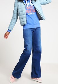 Giacca puffer azzurro chiaro sopra un hoodie blu con lacci bianchi, abbinata a jeans a zampa di elefante in denim scuro e sneakers rosa.