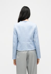 Γυναίκα με μακριά σκουρόχρωμα μαλλιά, φορόντας ανοιχτό μπλε υφασμάτινο σακάκι με ξεφτισμένες άκρες και γκρι παντελόνι με λεπτές ρίγες, φαίνεται από την πίσω πλευρά.