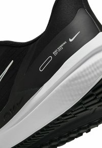 Zapatilla de correr negra con parte superior de tejido texturizado, suela de goma blanca y diseño minimalista. Presenta la marca Nike en blanco.