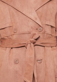 Palton din faux suede roz pal, cu talie evidențiată prin curea, față dublu-captusită și trei nasturi. Include guler și o textură distinctivă.