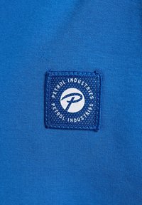 Fond en tissu bleu avec un patch rectangulaire comportant le texte "Petrol Industries" et un logo en blanc, surface texturée le long des bords.