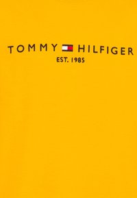 Camiseta de algodón amarilla con el logo de "TOMMY HILFIGER" en color navy, con un pequeño gráfico de bandera roja y blanca y "EST. 1985" debajo.
