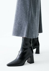 Stivaletti in pelle nera con punta appuntita e tacco largo. Gli stivaletti hanno una texture liscia e sono abbinati a pantaloni grigi in tessuto tagliati sopra la caviglia.