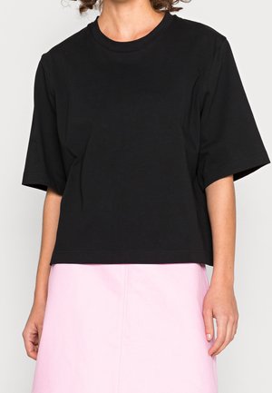 Person trägt ein lockeres schwarzes Kurzarm-T-Shirt und einen hellrosa Rock vor einfarbigem Hintergrund.