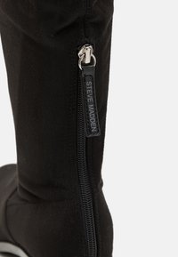 Black fabric knee-high boot featuring a sleek design, pointed toe, and silver zipper with branding along the side. Smooth texture.