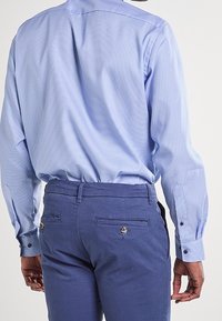 Uomo che indossa una camicia a maniche lunghe blu chiaro infilata in pantaloni blu navy, visto da dietro, in piedi contro uno sfondo semplice.