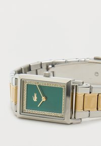 Roheline ristkülikukujuline kell hõbedasest ja kuldse tooni metallist rihmaga. Omab kristallidega äärist ja kuldset logo kellanäidiku peal. Kuldse värvusega kellanäidikud.