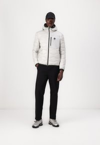 Giacca grigia imbottita con cappuccio e zip frontale, dotata di tasca sul petto. I pantaloni neri e le sneakers silver completano l'outfit.