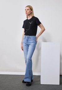 Svart T-skjorte med hvit linjekunstdesign, sammen med lyseblå bukser med vide ben. Modellen står mot en nøytral bakgrunn. Svart platåsko.