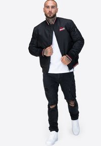 Czarna kurtka bomberka z czerwono-białym ściągaczem, z zamkiem z przodu i logo. Noszona na białej koszulce, zestawiona z przetartymi czarnymi dżinsami i białymi sneakersami.