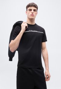 Νεαρός άνδρας που φοράει μαύρο t-shirt Calvin Klein και μαύρο παντελόνι, κρατώντας ένα μαύρο σακάκι ακουμπισμένο στον ώμο του, μπροστά σε απλό φόντο.