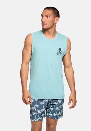 Hombre con camiseta sin mangas azul claro con logotipo de palmera y pantalones cortos azul marino con estampado de palmeras blancas, mirando hacia un lado.