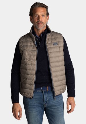 Beige gewatteerde vest met een staande kraag, ritssluiting en een marineblauwe trui eronder. Opvallende textuurpatronen op de trui. Blauwe spijkerbroek met een riem.