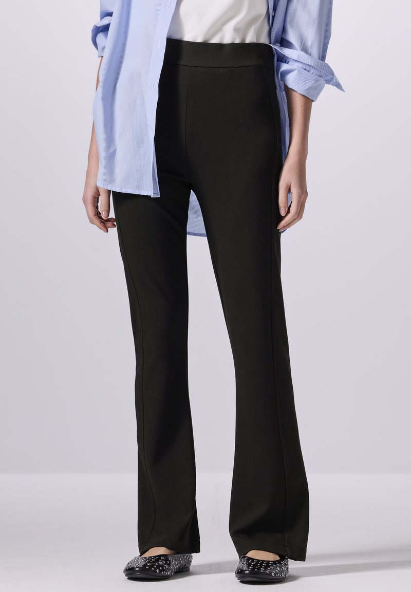 Pantaloni neri a zampa d'elefante realizzati in tessuto elastico, caratterizzati da una vita alta e una texture liscia, abbinati a una camicia azzurra con colletto.