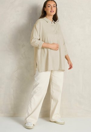 Kvinde står op ad en neutral væg iført en beige oversized skjorte med knapper, hvide bukser med vide ben og hvide sneakers.