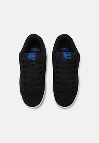 Baskets noires avec une finition texturée, présentant un design bas, des lacets plats et une semelle en caoutchouc blanche. Accents de logo bleu sur la languette.