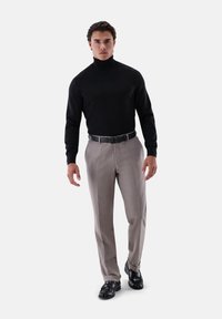 Mann in schwarzem Rollkragenpullover, beigen Hosen mit schwarzem Gürtel und schwarzen Lederschuhen steht vor weißem Hintergrund.