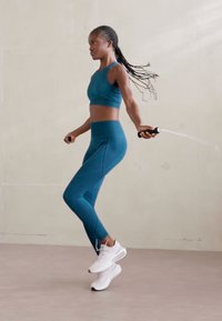 Jogador atlético en tejido azul verdoso, con top corto y leggings a juego; lleva zapatillas blancas, cuerda de saltar y tiene el cabello trenzado en movimiento.