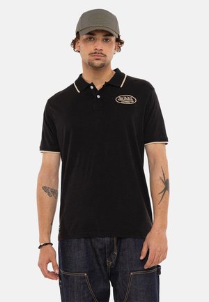 Homme portant un polo noir Von Dutch avec des bordures beiges, une casquette de baseball grise Von Dutch, et un jean foncé, montrant des tatouages de papillon et d'araignée sur les bras.