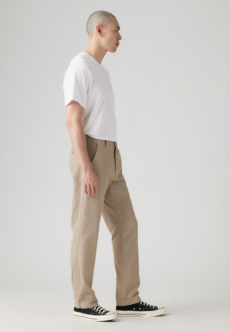 Chinos beiges, en coton, coupe droite, associés à un t-shirt blanc à manches courtes et des baskets noires avec des semelles blanches.