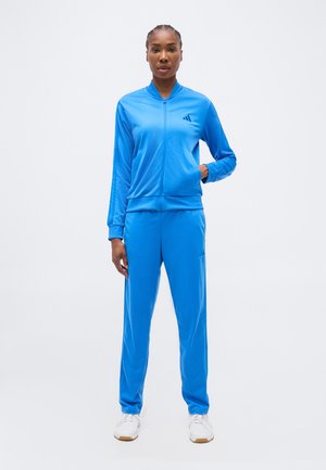 Kobieta stoi, ubrana w jaskrawo niebieski dres z kurtką na zamek i dopasowanymi spodniami, do tego białe trampki, na jednolitym tle.