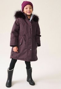 Lilla parka med faux fur kant på hætten, trykknap- og lynlåslukninger, to frontlommer, kombineret med sorte leggings og gummistøvler.