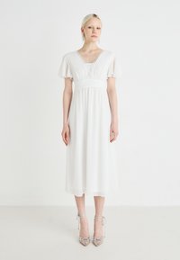 Valge midi kleit lühikeste lipukeste varrukatega, volditud rinnaosaga ja V-kaelusega. Mudel kannab kaunistatud kõrgeid kontsi.