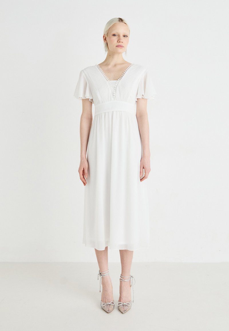 Valge midi kleit lühikeste lipukeste varrukatega, volditud rinnaosaga ja V-kaelusega. Mudel kannab kaunistatud kõrgeid kontsi.