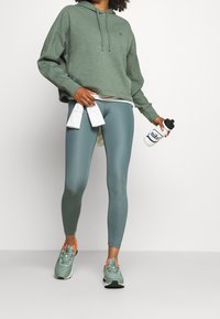 Felpa verde, leggings azzurri e sneakers verdi con dettagli arancioni. Tiene in mano una borraccia bianca e un asciugamano bianco.