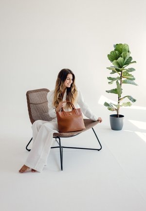 Braune Leder-Tote mit kurzen Griffen, mit einer glatten Textur und kleinem Logo. Im Hintergrund sind ein geflochtener Stuhl und eine grüne Pflanze.