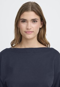Fiatal nő, világosbarna hajjal, sötét tengerészkék, hajónyakú pulóvert visel, egyenesen előttére nézve, semleges arckifejezéssel.