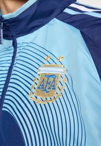 Ανοιχτό μπλε αθλητικό σακάκι με ναυτικά στοιχεία, με οριζόντιες ρίγες. Διαθέτει κεντητό χρυσό λογότυπο με δάφνινο στεφάνι και αστέρια.