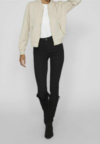 Personne portant une veste bomber beige, une chemise blanche, un jean skinny noir taille haute et des bottes noires mi-mollet, debout devant un fond uni.