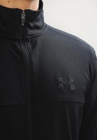 Černá bunda na zip z texturované látky, s vysokým límcem a vyšitým logem na levé straně hrudi.