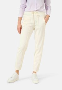 Kobieta ubrana w kremowe spodnie z troczkami i bocznymi kieszeniami, w białych trampkach na platformie, stojąca na tle białym.