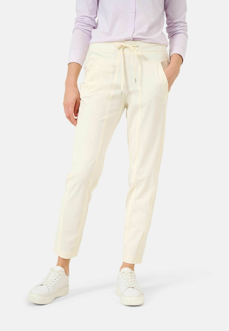 Kobieta ubrana w kremowe spodnie z troczkami i bocznymi kieszeniami, w białych trampkach na platformie, stojąca na tle białym.