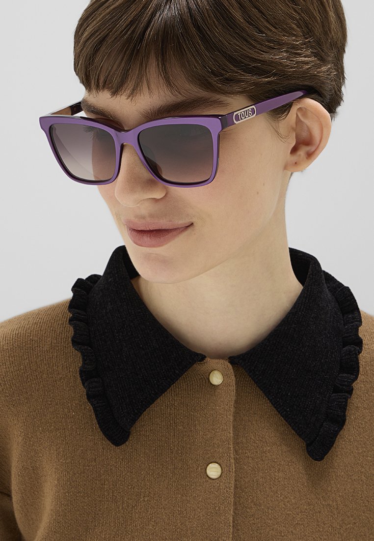 Fioletowe prostokątne okulary przeciwsłoneczne z gradientowymi soczewkami, noszone z brązowym swetrem z czarnym falbanowanym kołnierzykiem i zapięciem na guziki.
